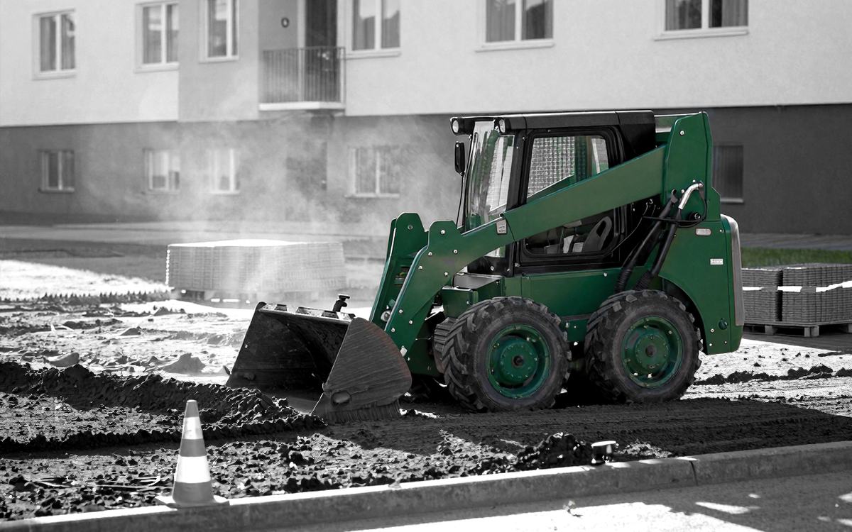 Мини-погрузчик разравнивает грунт на площадке во дворе: рычаги подъёма идут по бокам корпуса, ковш опущен на землю